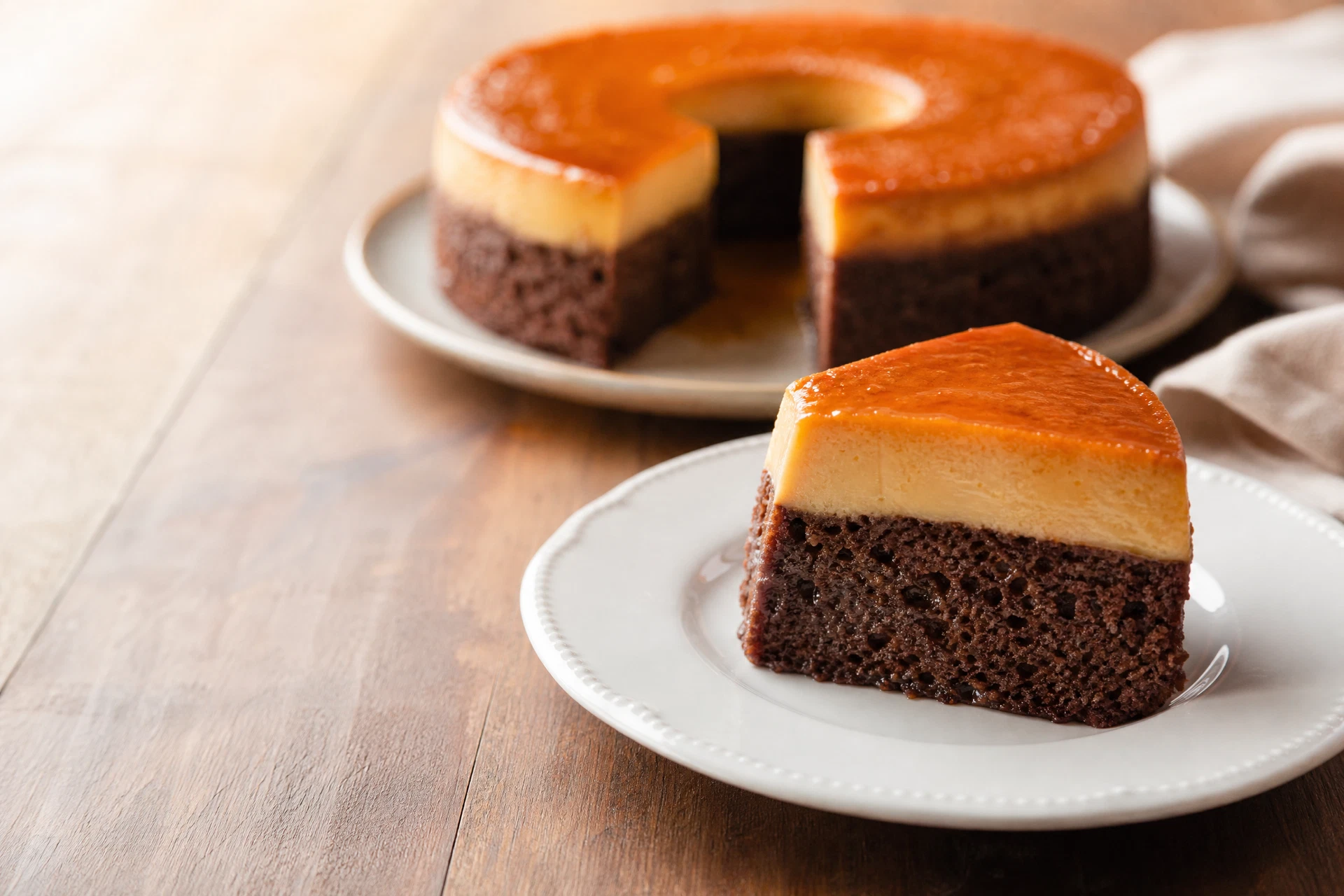 Fatia de bolo pudim com camada de chocolate, pudim claro e calda de caramelo.
