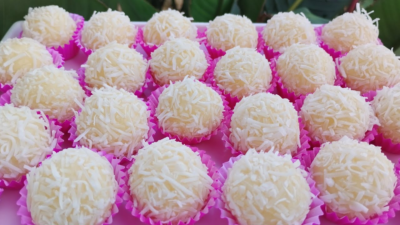 Fotografia em close de brigadeiros de beijinho cremosos cobertos com coco ralado e decorados com cravo-da-índia, dispostos em forminhas de papel sobre uma mesa de festa rústica.