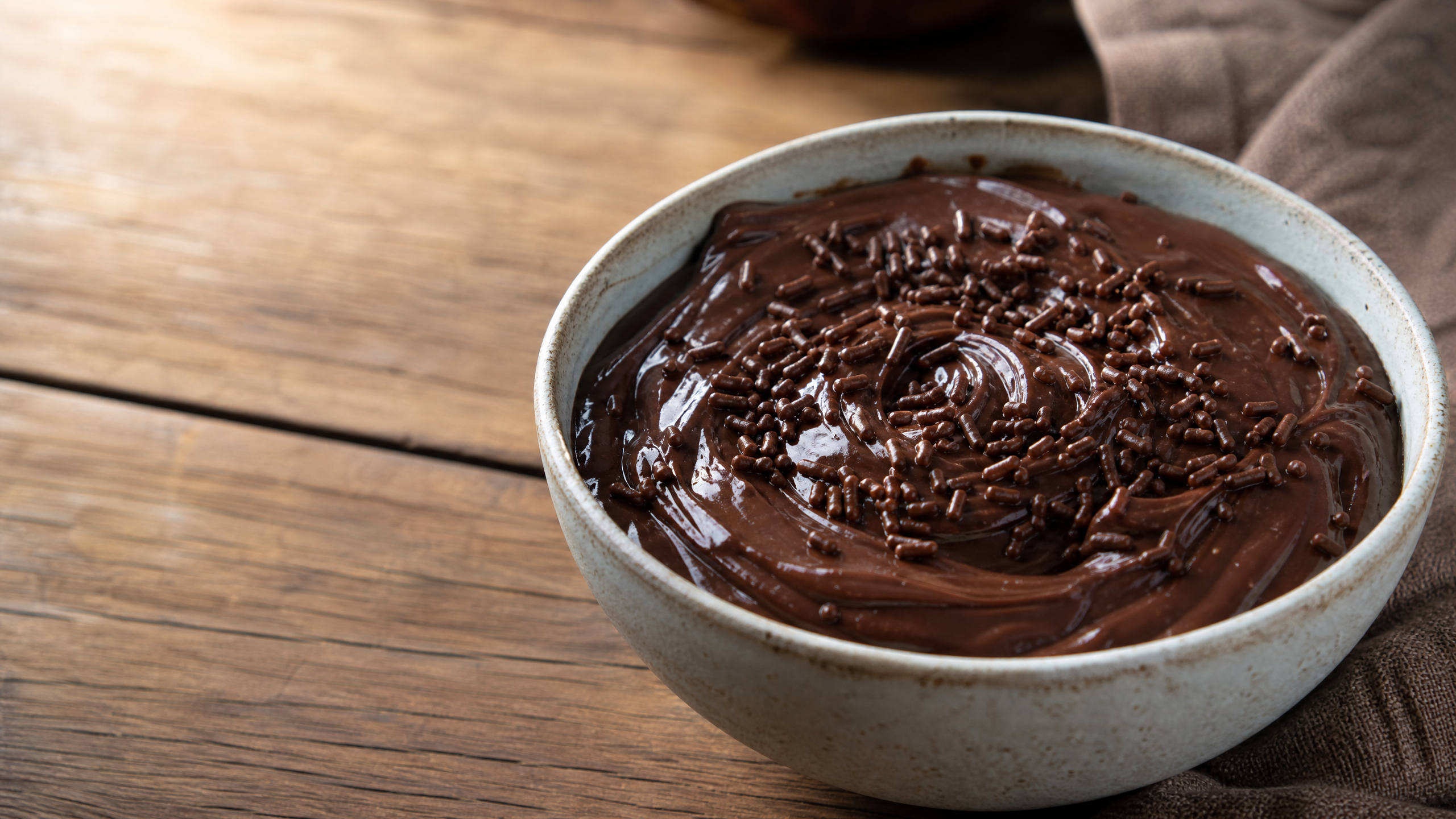 Prato raso branco com porção generosa de brigadeiro cremoso e brilhante, finalizado com granulado de chocolate e acompanhado por uma colher, visto de cima em superfície de madeira rústica.
