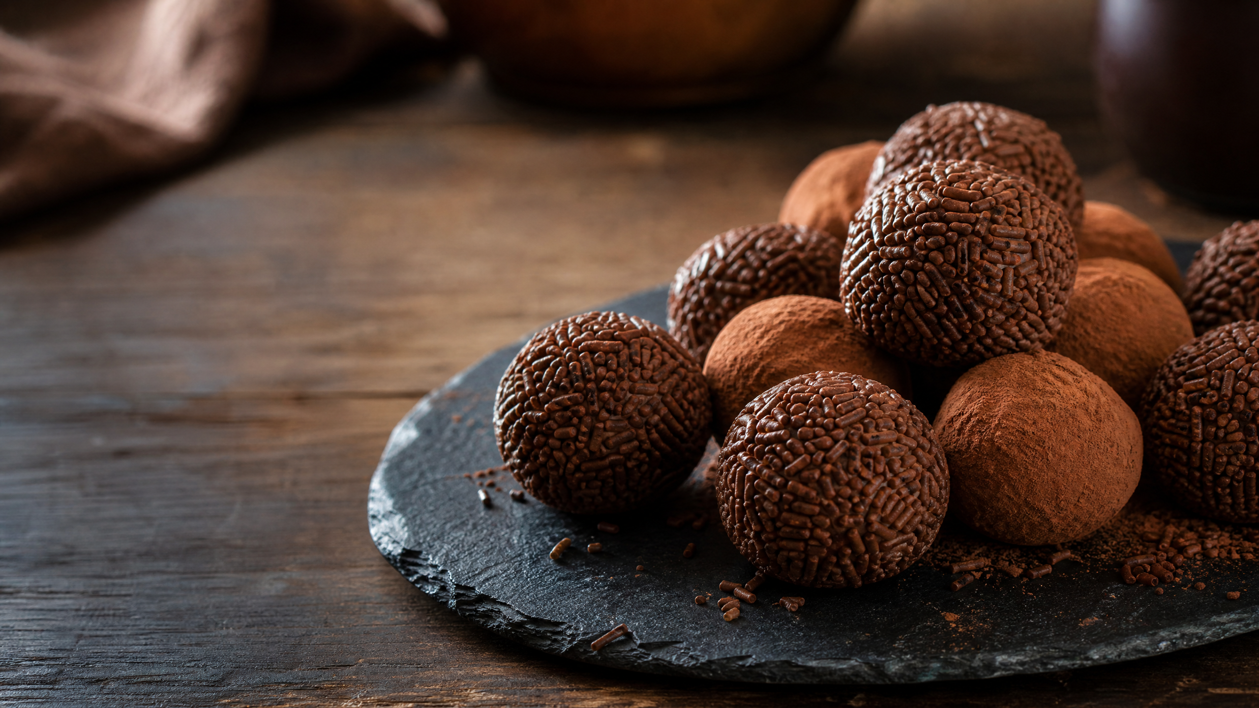 Brigadeiros gourmet enrolados artesanalmente dispostos em forminhas elegantes, cobertos com confeitos de chocolate granulado nobre e fundo desfocado mostrando utensílios de confeitaria.