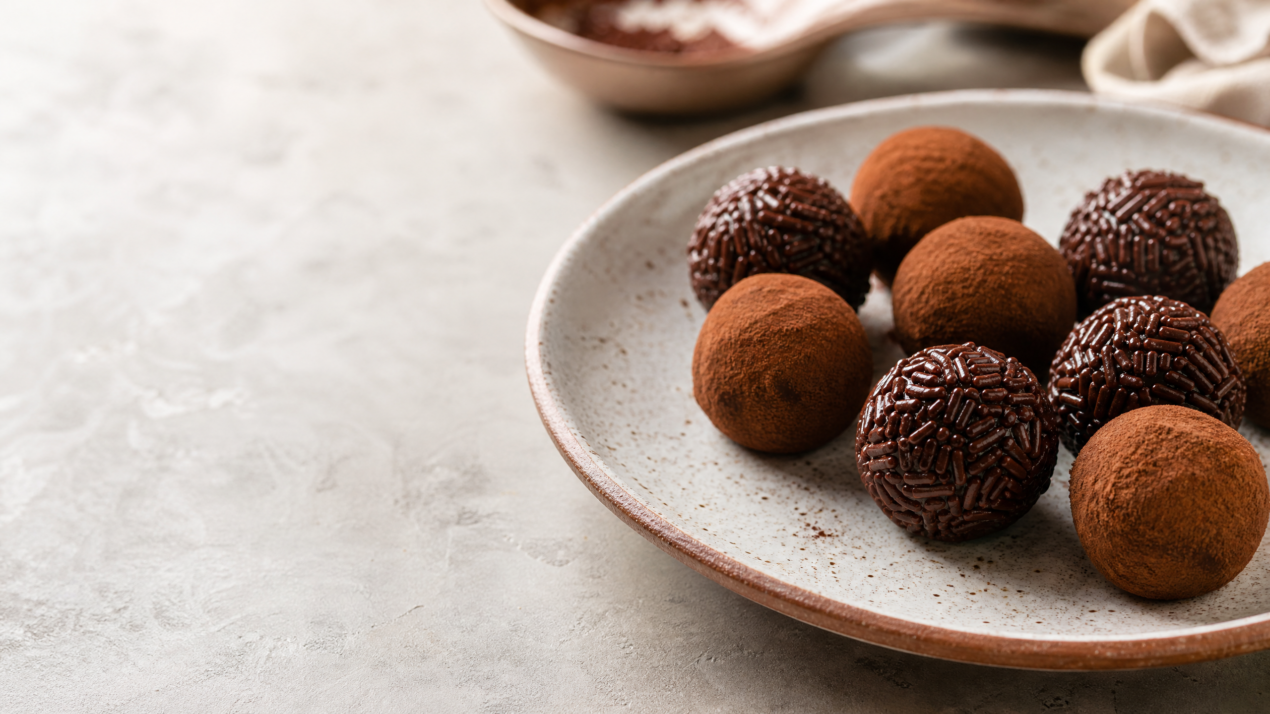 Brigadeiro vegano cremoso em formato de colherada, polvilhado com cacau em pó sobre fundo neutro de madeira escura, destacando textura brilhante e homogênea.