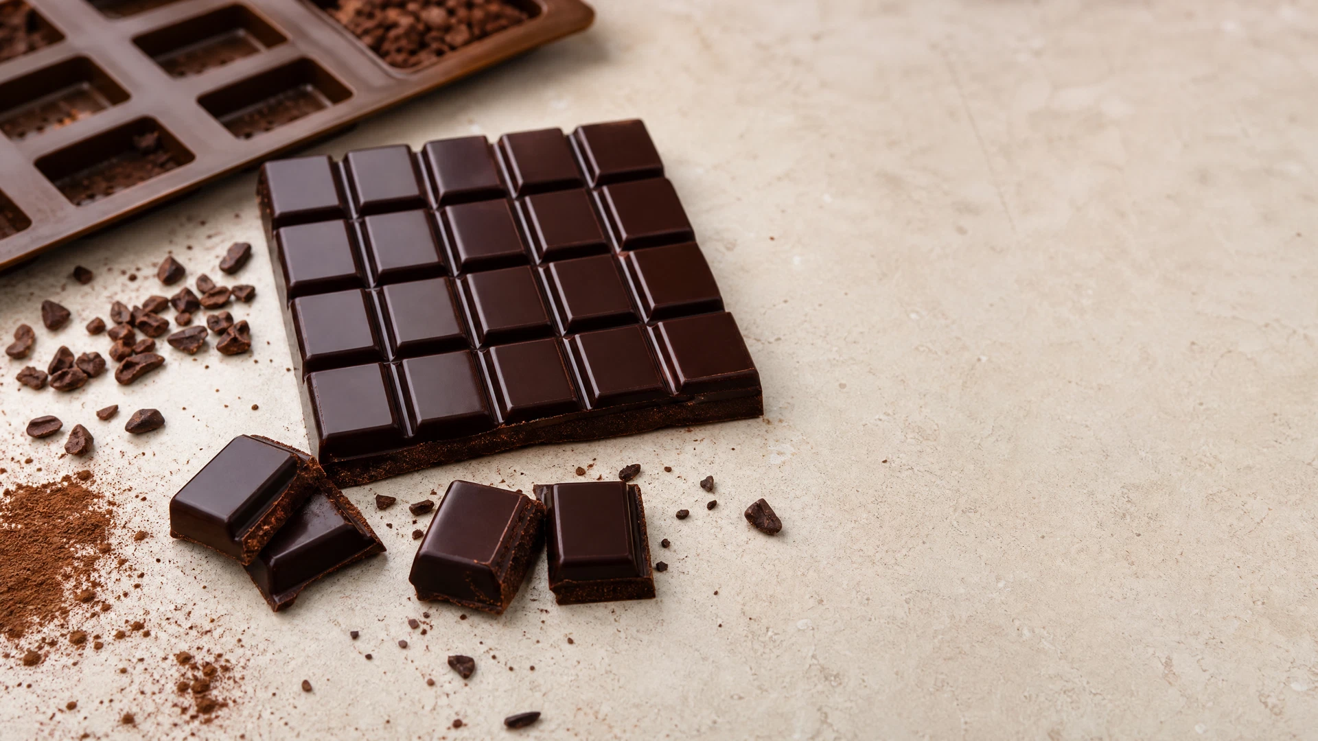 Barra de chocolate caseiro moldada em quadrados, com cacau em pó e nibs de cacau sobre superfície clara.