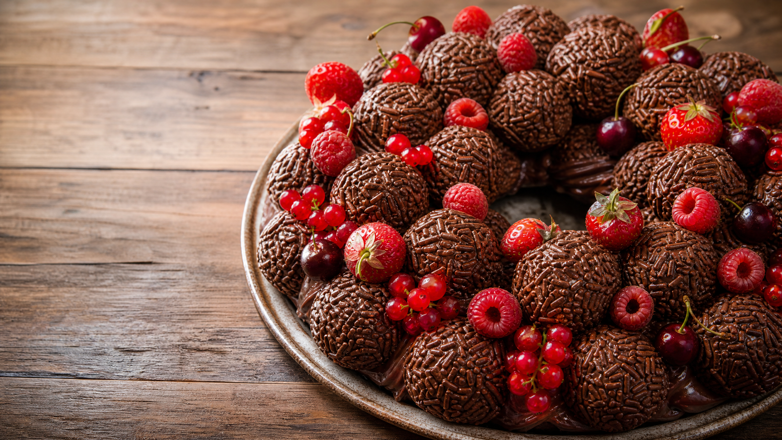 Vista superior de uma guirlanda de brigadeiro artesanal montada em base circular, decorada com brigadeiros enrolados em granulado escuro e confeitos perolados, dispostos sobre uma mesa com detalhes festivos.