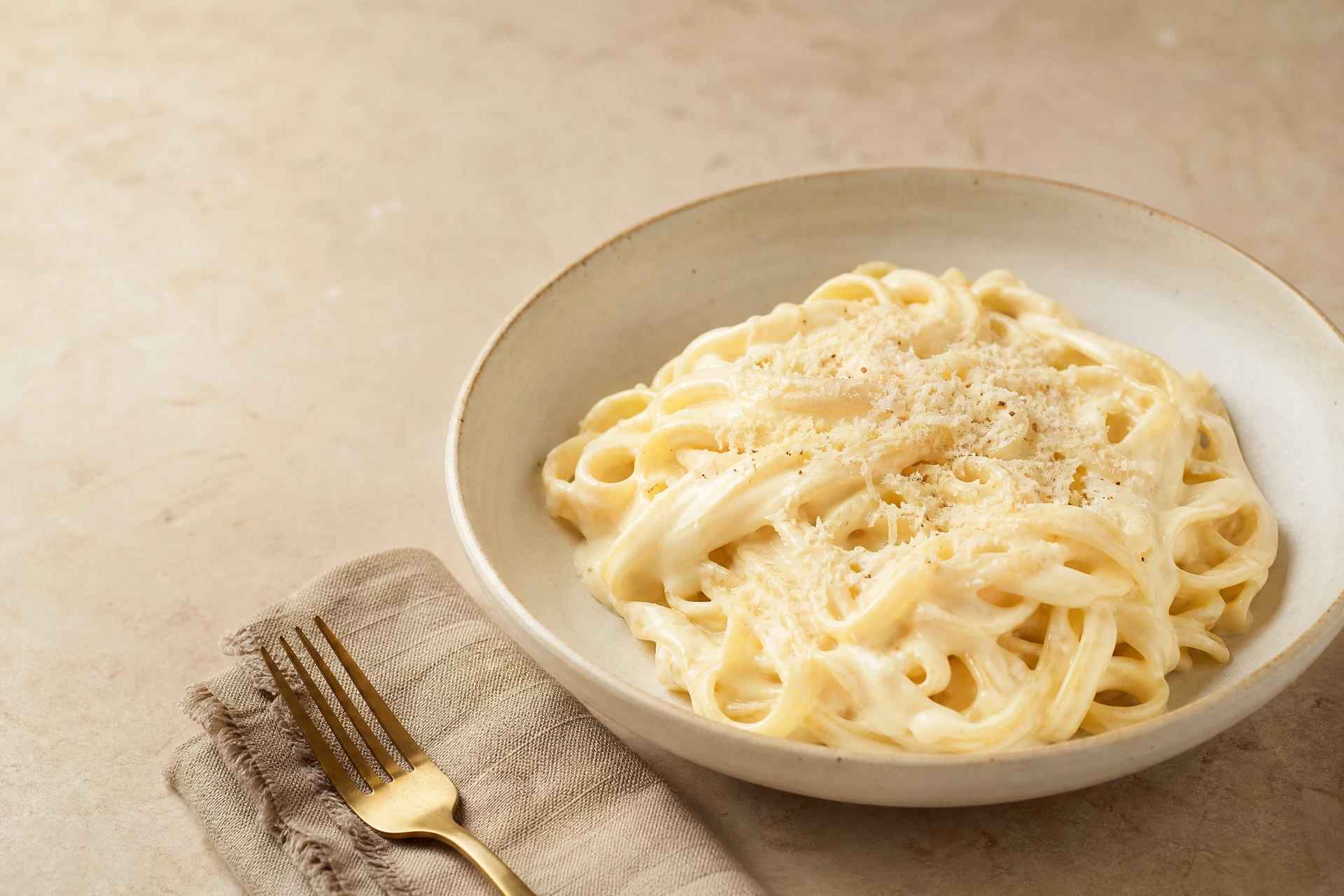 Tigela de fettuccine envolvido por molho branco cremoso sobre mesa clara, com parmesão ralado fino por cima.