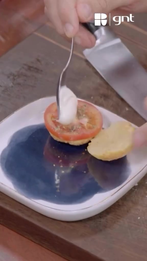 Creme de ricota sendo colocado sobre tomate e orégano no pão de queijo aberto, com marca d'água GNT preservada.