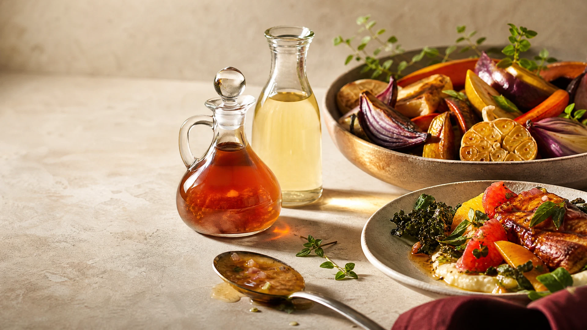 Garrafas sem rótulo de vinagre e vinagrete ao lado de vegetais assados em mesa de pedra clara.